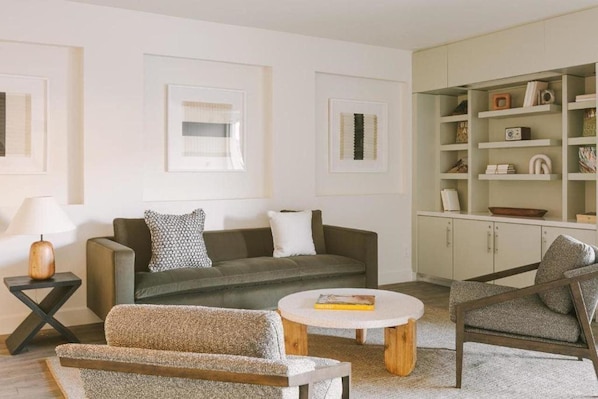 Una sala moderna con un sofá, un sillón y una mesa de centro. Se aprecia una estantería con diversos objetos y fotografías enmarcadas en la pared.