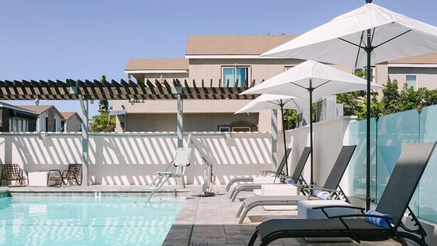 Ein Poolbereich mit Liegestühlen und Sonnenschirmen, einer Pergola sowie ein Gebäude mit einem Ziegeldach im Hintergrund.
