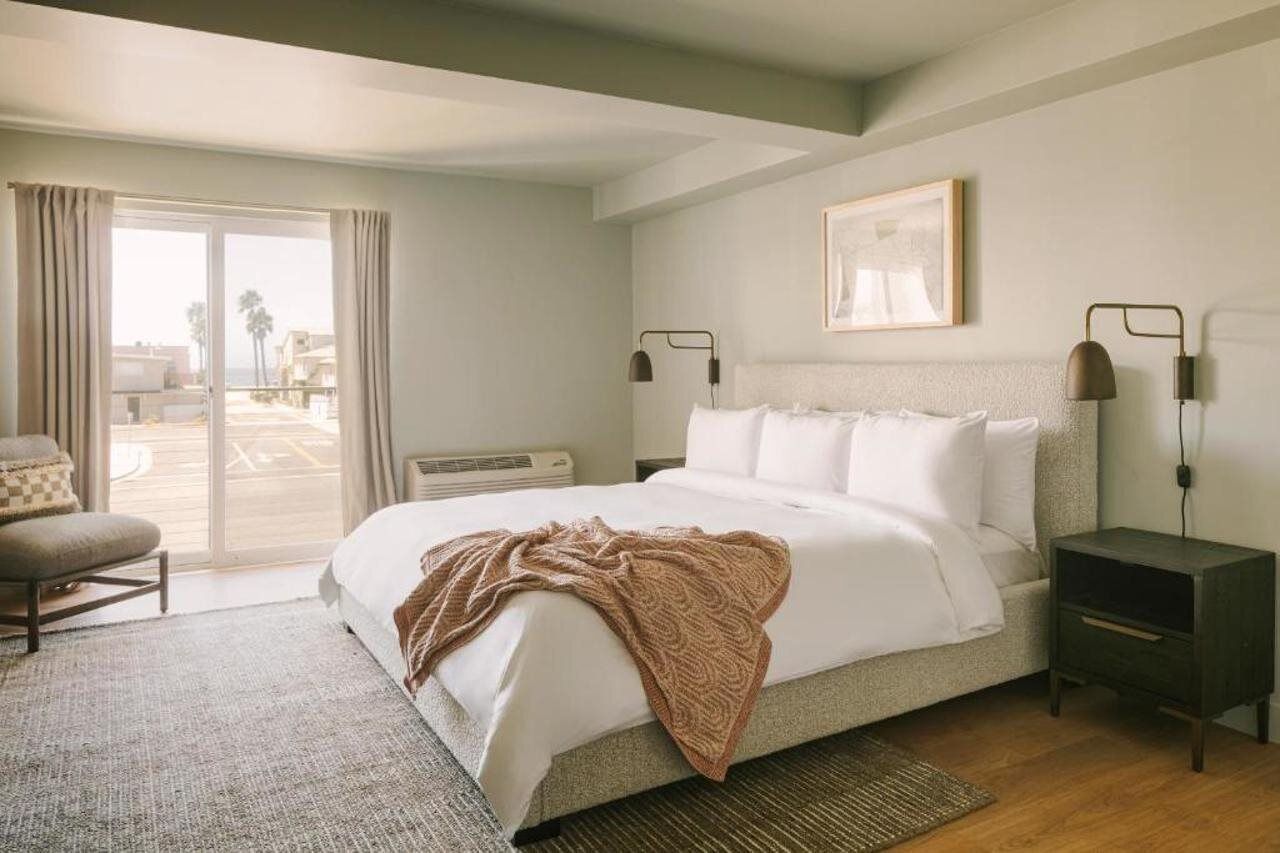 Un dormitorio con cama, una silla, una mesita de noche, una lámpara y una ventana con cortinas.
