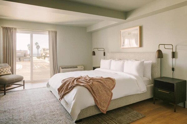 Un dormitorio con cama, una silla, una mesita de noche, una lámpara y una ventana con cortinas.