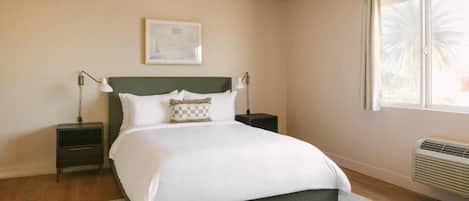 Un dormitorio con cama, dos mesitas de noche, una mesilla, una lámpara de pared, un cuadro enmarcado, una ventana con cortinas y un radiador.