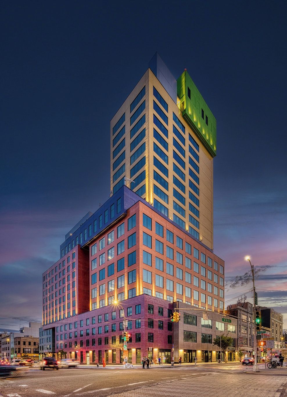 A multi-story hotel with a prominent green sign on top, a large number of windows, and a street view with traffic lights and pedestrians.