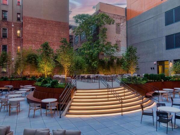An outdoor patio area with tiered seating, tables, and chairs, surrounded by greenery and buildings. - Radio Hotel (New York)