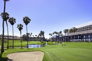 A golf course with a pond, palm trees, and a modern building in the background.