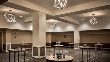 A spacious hotel lobby with geometric ceiling lights, multiple tables with plants, and a carpeted floor.