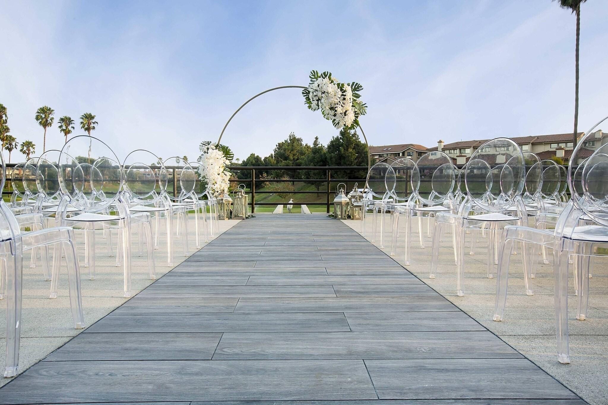 On peut clairement voir un chemin en bois qui mène à un espace aménagé avec des chaises transparentes, une arche fleurie et, en arrière-plan, des palmiers et des bâtiments.