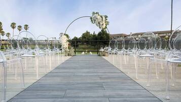 On peut clairement voir un chemin en bois qui mène à un espace aménagé avec des chaises transparentes, une arche fleurie et, en arrière-plan, des palmiers et des bâtiments.