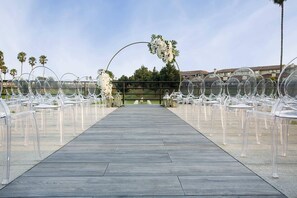 A clear view of a wooden pathway leading to a seating area with transparent chairs, a floral arch, and a backdrop of palm trees and buildings.