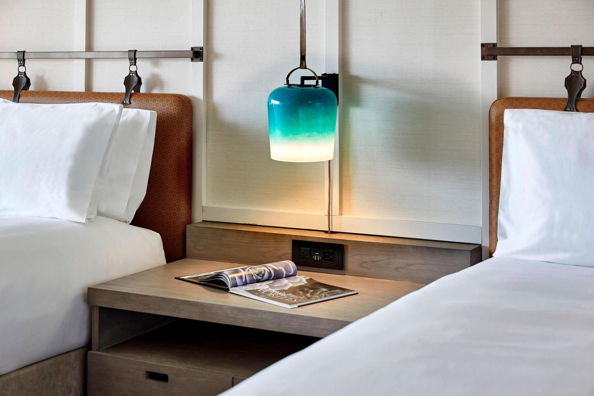 Une chambre d’hôtel avec deux lits, une table de chevet avec un magazine et une lampe fixée au mur.