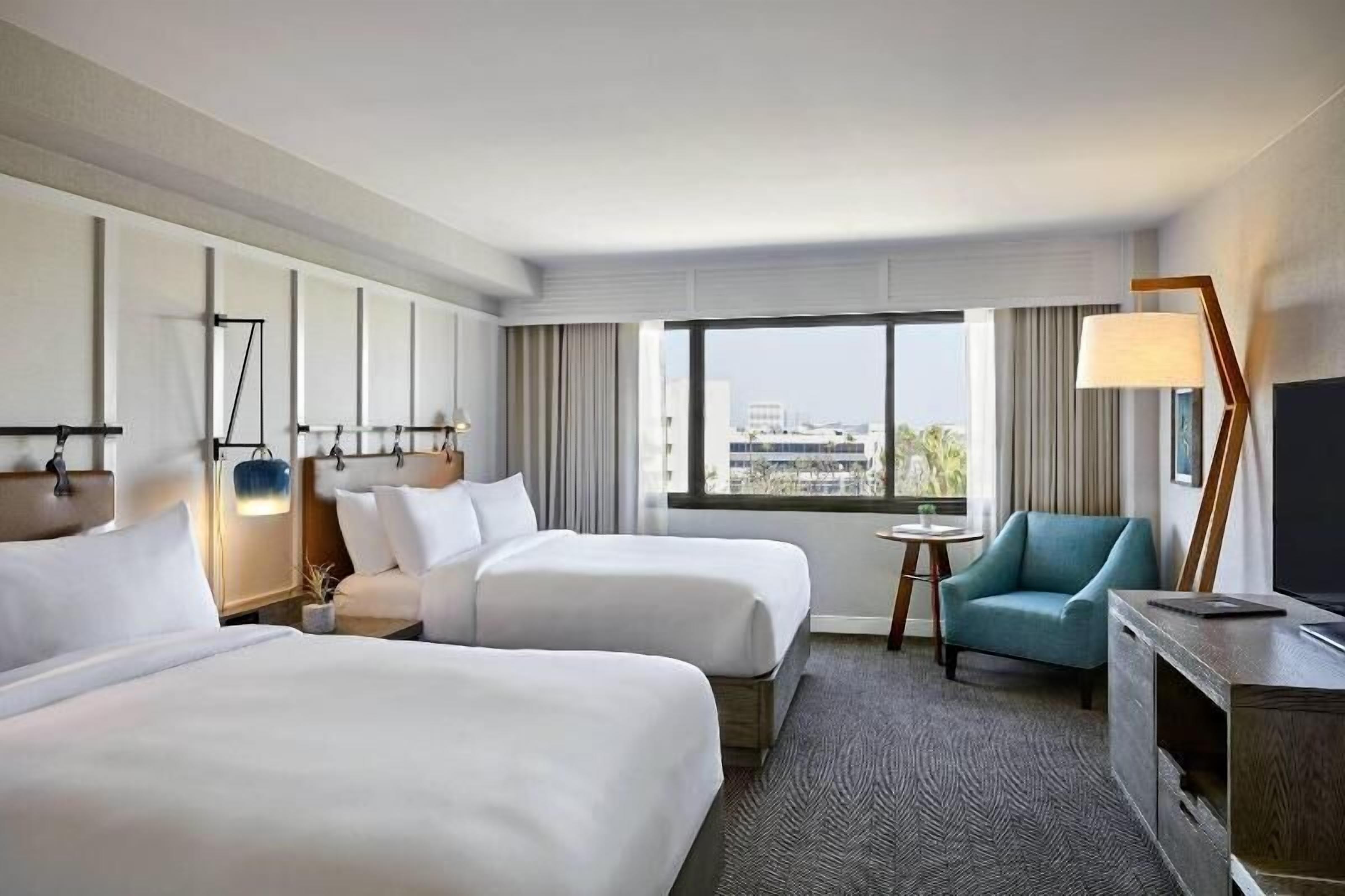 A hotel room with two beds, a desk, a chair, a TV, and a large window with a city view.