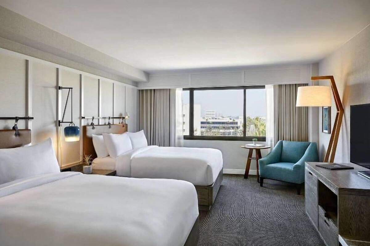 A hotel room with two beds, a desk, a chair, a TV, and a large window with a city view.
