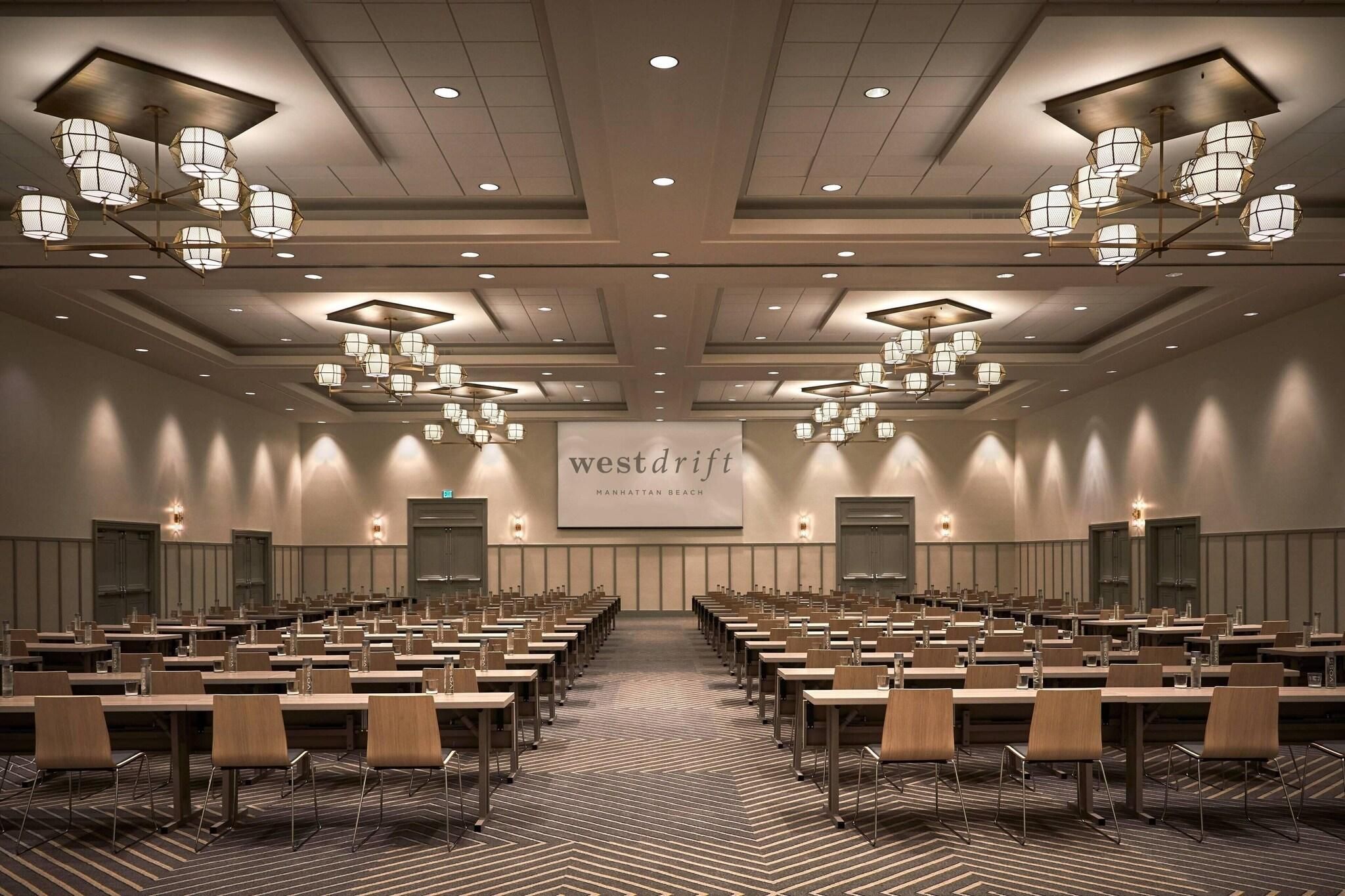 A conference room with rows of tables and chairs, a large screen, and chandeliers.