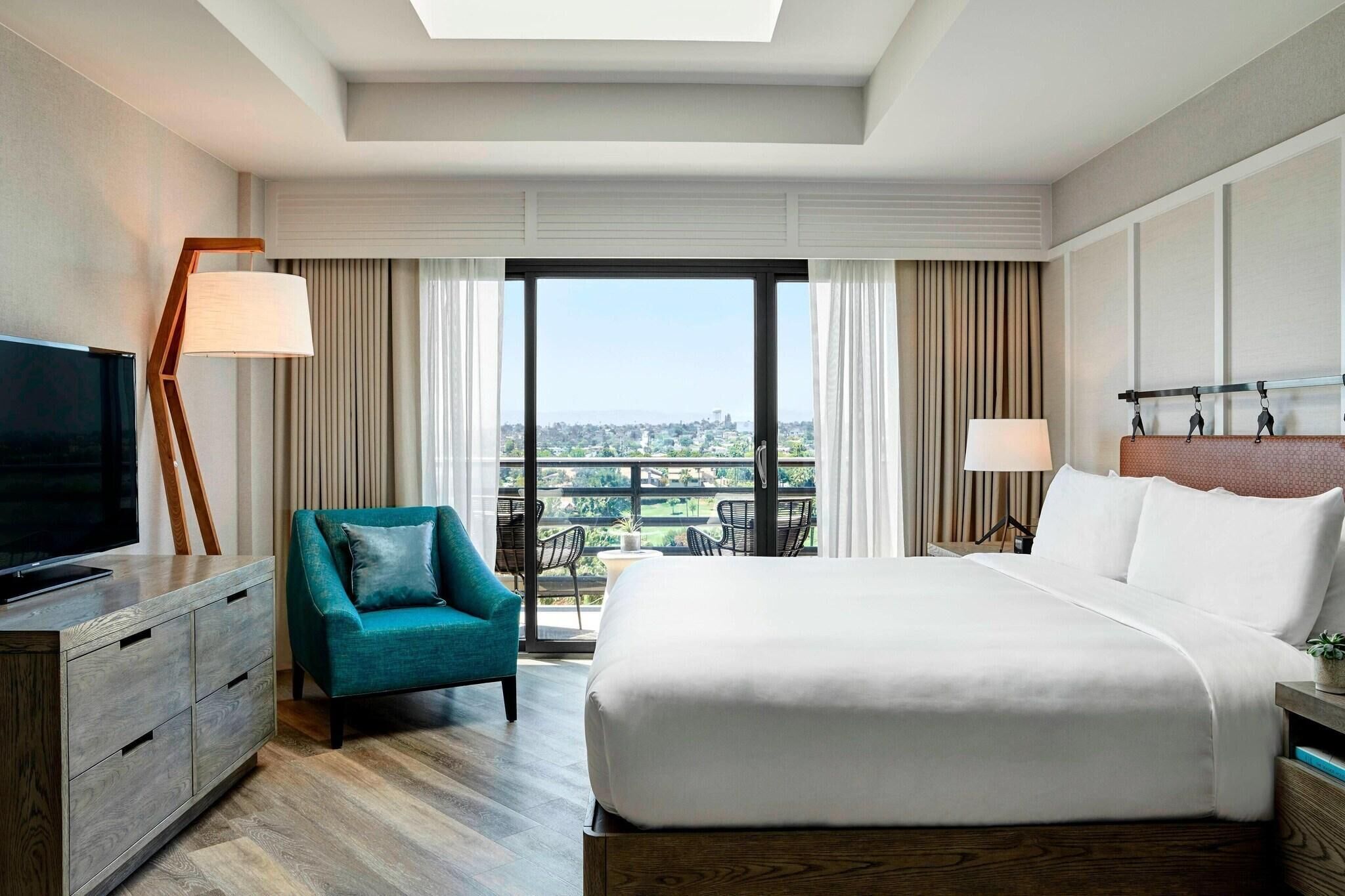 A hotel room with a bed, a TV, a chair, a lamp, and a view of the city.