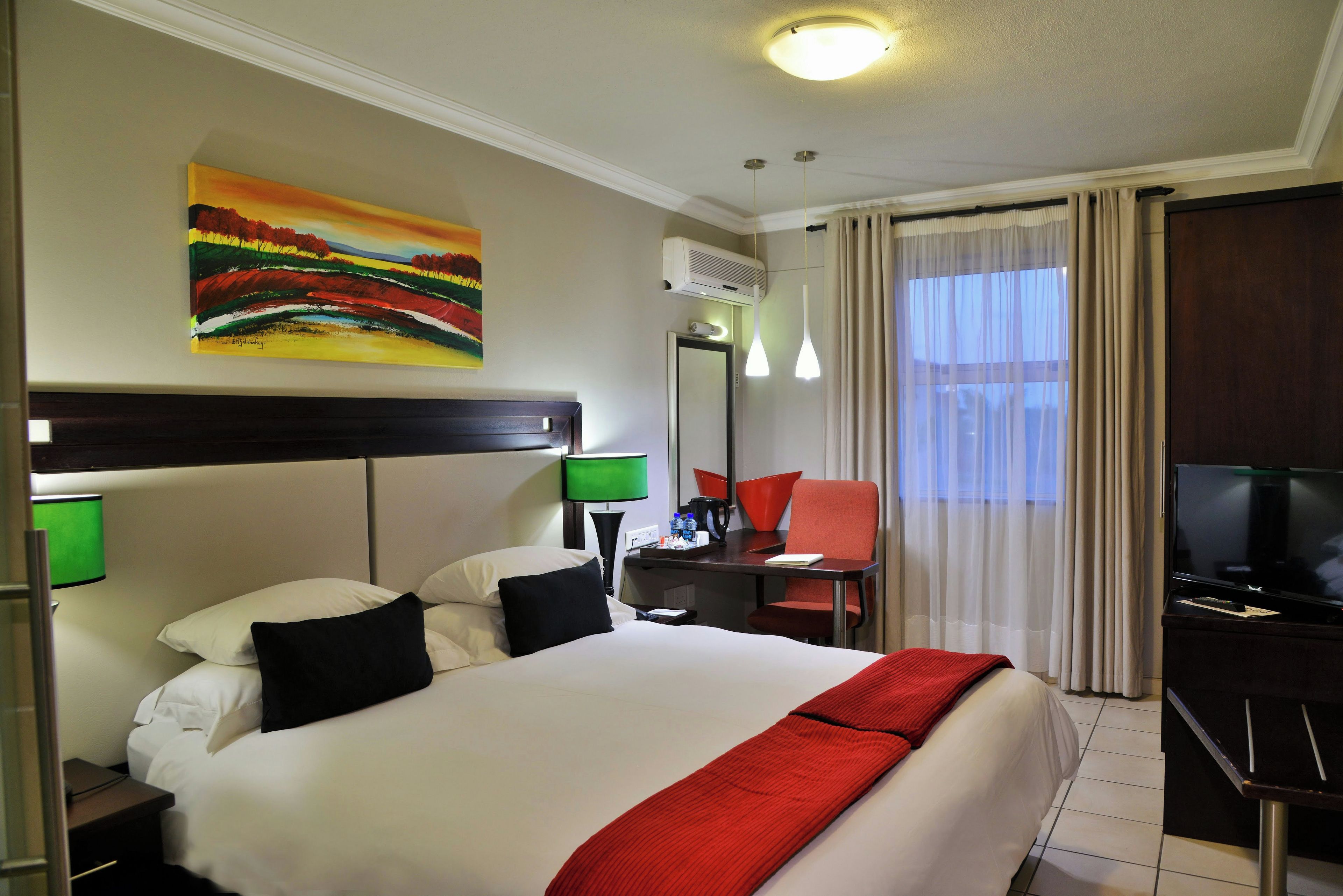 A hotel room with a bed, a desk, a chair, a television, and a painting on the wall.