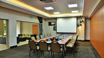 Un salón de conferencias con una larga mesa, sillas, una pantalla de proyector y una zona de estar con un sofá y cojines.
