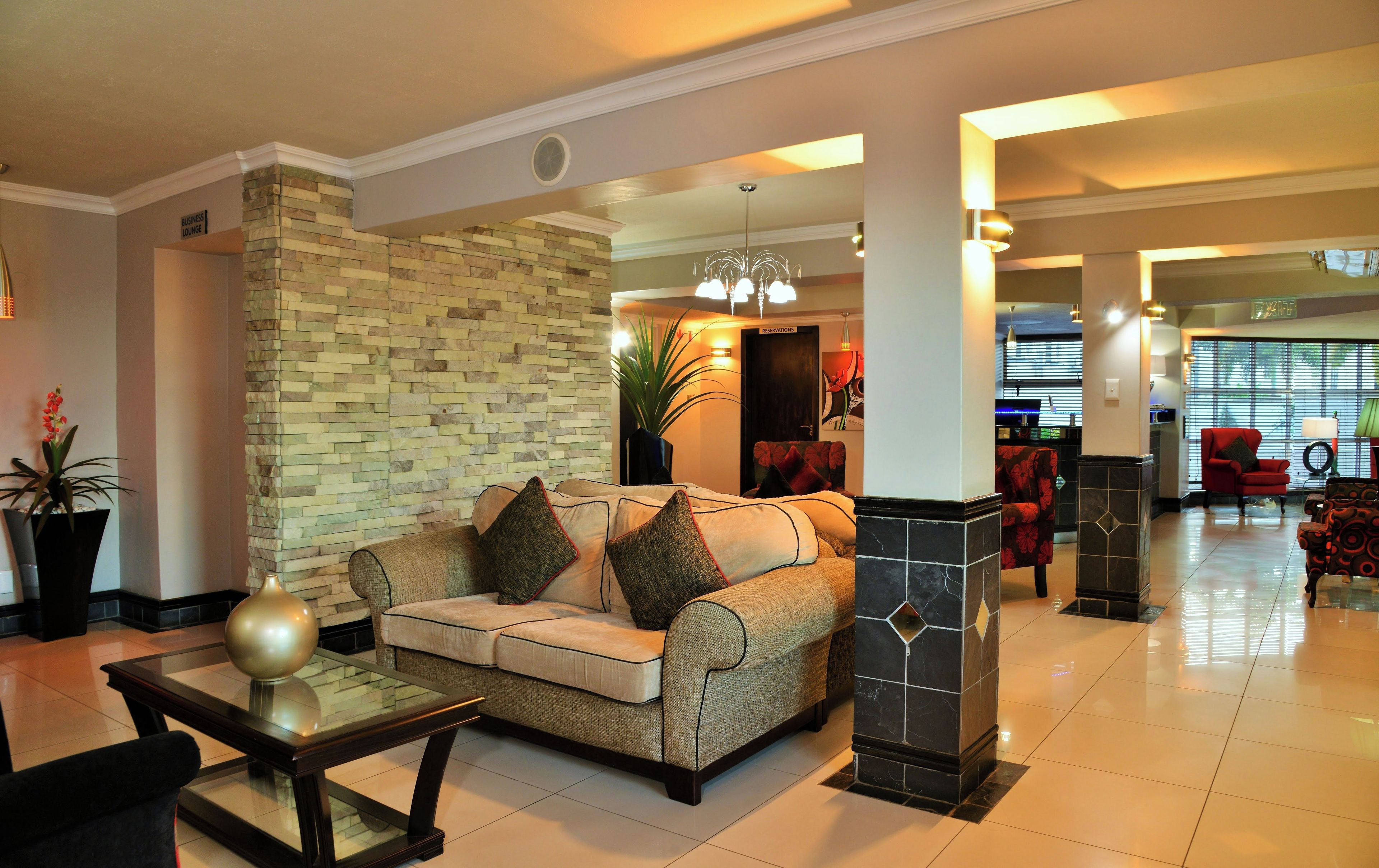 A hotel lobby with a sofa, a glass coffee table, a stone accent wall, a red armchair, and a reception desk.