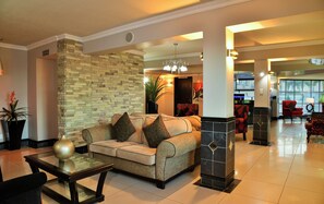A hotel lobby with a sofa, a glass coffee table, a stone accent wall, a red armchair, and a reception desk. - Bon Hotel Empangeni (Empangeni)