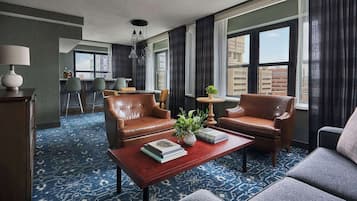 Habitación de hotel con sofá, sillones, una mesa de centro, lámpara y vistas de la ciudad a través de amplios ventanales.