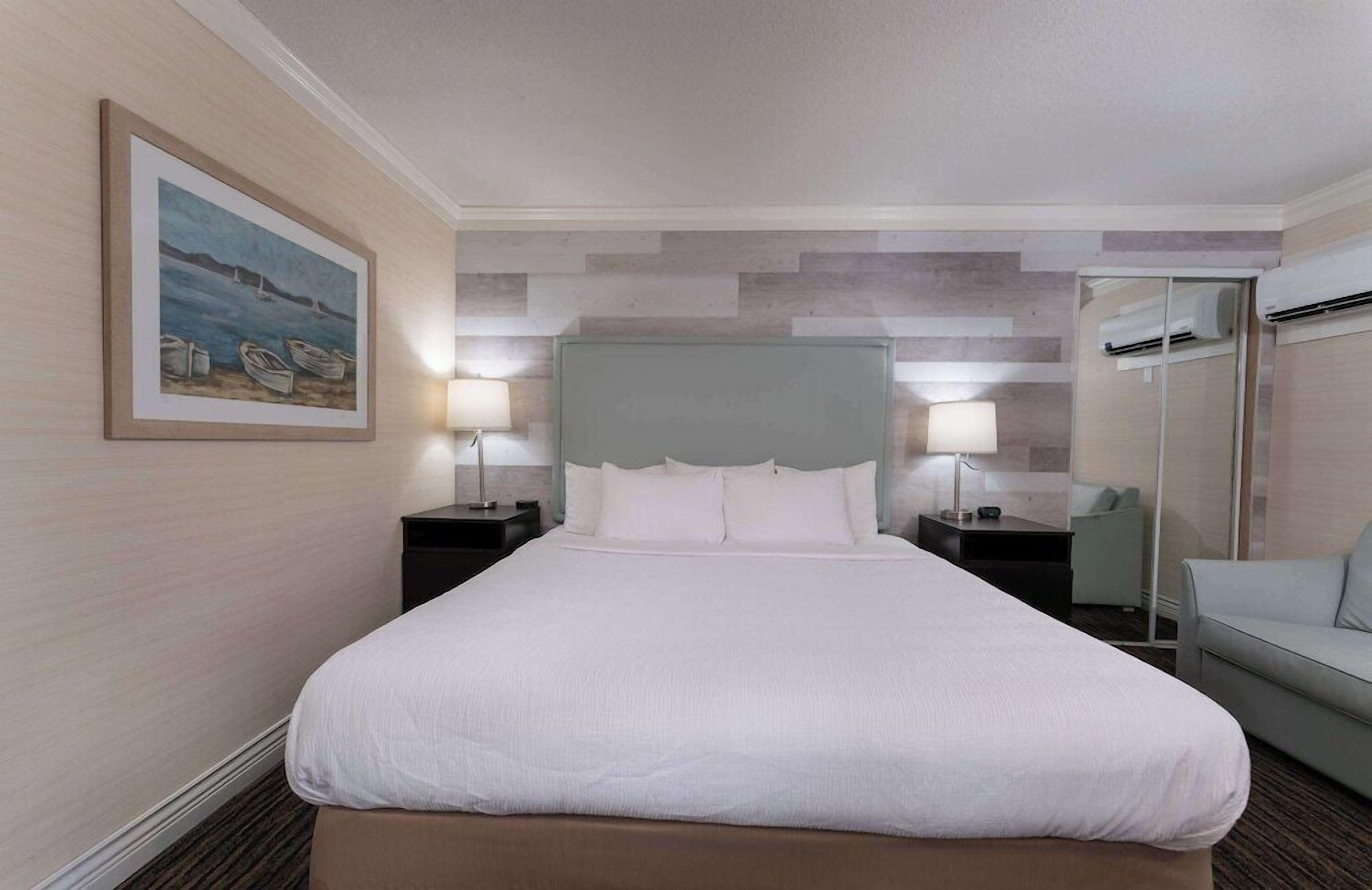 A hotel room with a large bed, two bedside tables with lamps, a grey headboard, a framed painting on the wall, and a grey sofa.