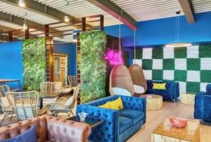 Un área de estar moderna con un sofá azul, sillas de mimbre y una pared verde con un letrero de neón.
