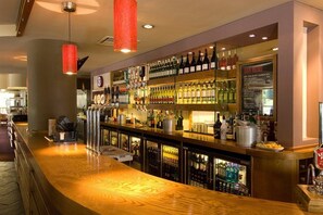 A bar area with a wooden counter, shelves of various liquor bottles, and a menu board. - Premier Inn Ashford (Ashford)