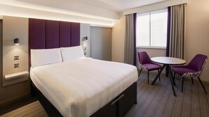 Una habitación de hotel con una cama grande, una mesita de noche, una lámpara, una silla, una pequeña mesa y una ventana con cortinas.