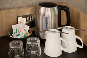Una encimera de cocina con un hervidor de acero inoxidable, dos tazas blancas, un tarro de vidrio y paquetes de té PG.