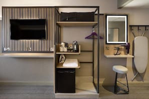 Une chambre d’hôtel compacte équipée d’une télévision, d’un petit lavabo, d’un miroir, d’un tabouret et d’un meuble de rangement avec un espace pour suspendre des vêtements.
