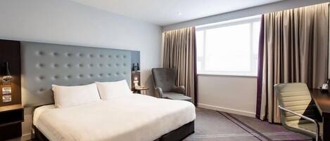 Quarto de hotel com uma cama grande, uma escrivaninha, uma cadeira e uma janela com cortinas.