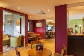 A modern hotel lobby with a wooden reception desk, a dining area with a round table and chairs, and a seating area with sofas and armchairs. - Premier Inn Worcester City Centre (Worcester)