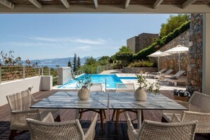 Un espace aménagé au bord de la piscine, avec une table et des chaises, un mur en pierre et une vue sur la mer et les montagnes.