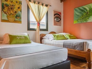 A hotel room with two beds, each with a green pillow labeled "Ayenda HOTEL." There is a window with a curtain, a painting of sunflowers, and another painting of an eye on the wall. - Habana Vieja Hotel (Medellín)
