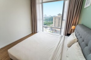 Quarto de hotel com uma janela grande, uma cama com roupa de cama branca, cabeceira cinzenta e uma luminária de cabeceira.