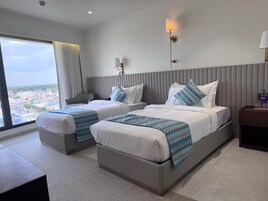 A hotel room with two beds, a large window with a city view, and a nightstand with a lamp.