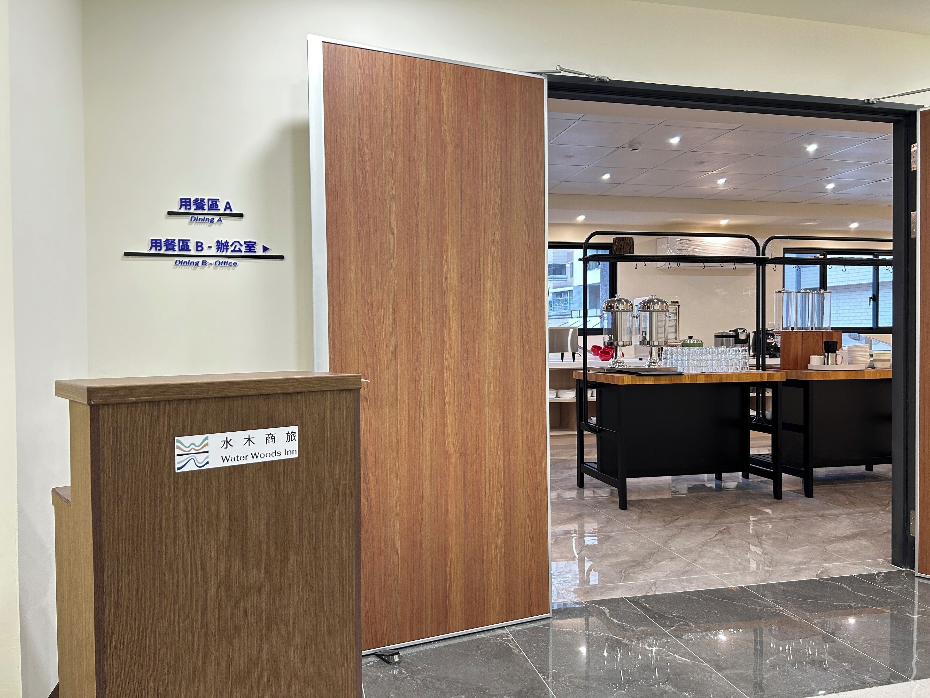 Een hotel receptie met een houten balie, een bord met de naam Water Woods Inn, en een glimp van de ontbijtruimte op de achtergrond.
