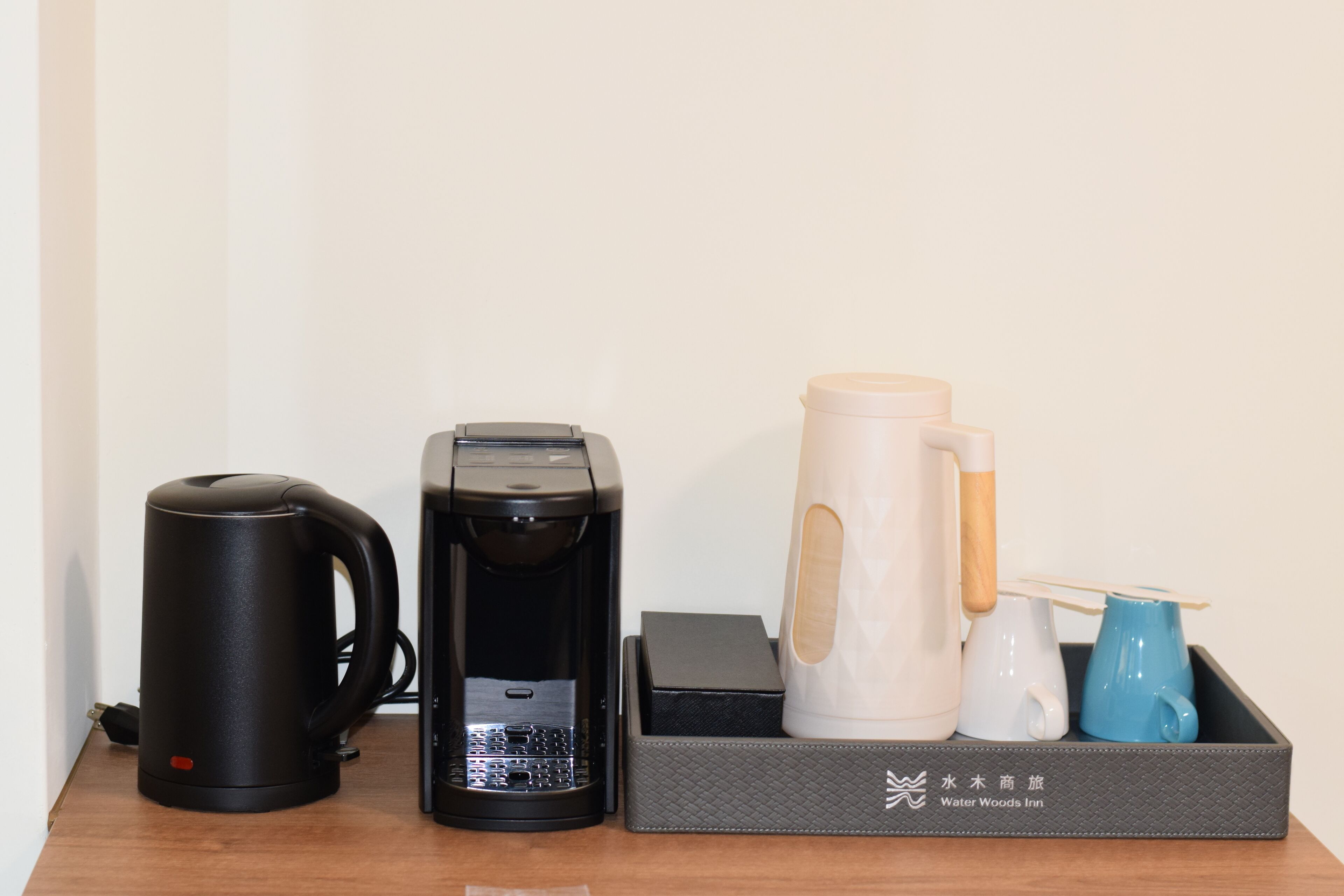 A coffee maker, a water kettle, and a tray with a water jug and cups on a wooden table.