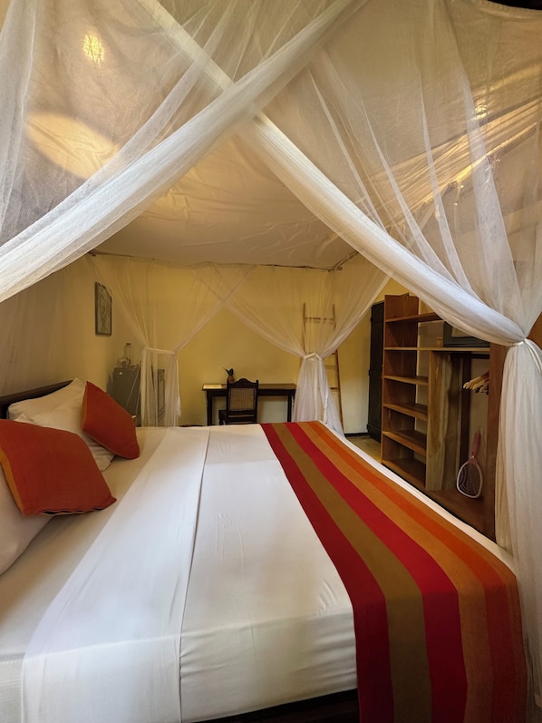 A bedroom with a bed, pillows, a bedside table, a chair, a wardrobe, and a canopy bedspread. - Dunes Unawatuna Hotel (Unawatuna)