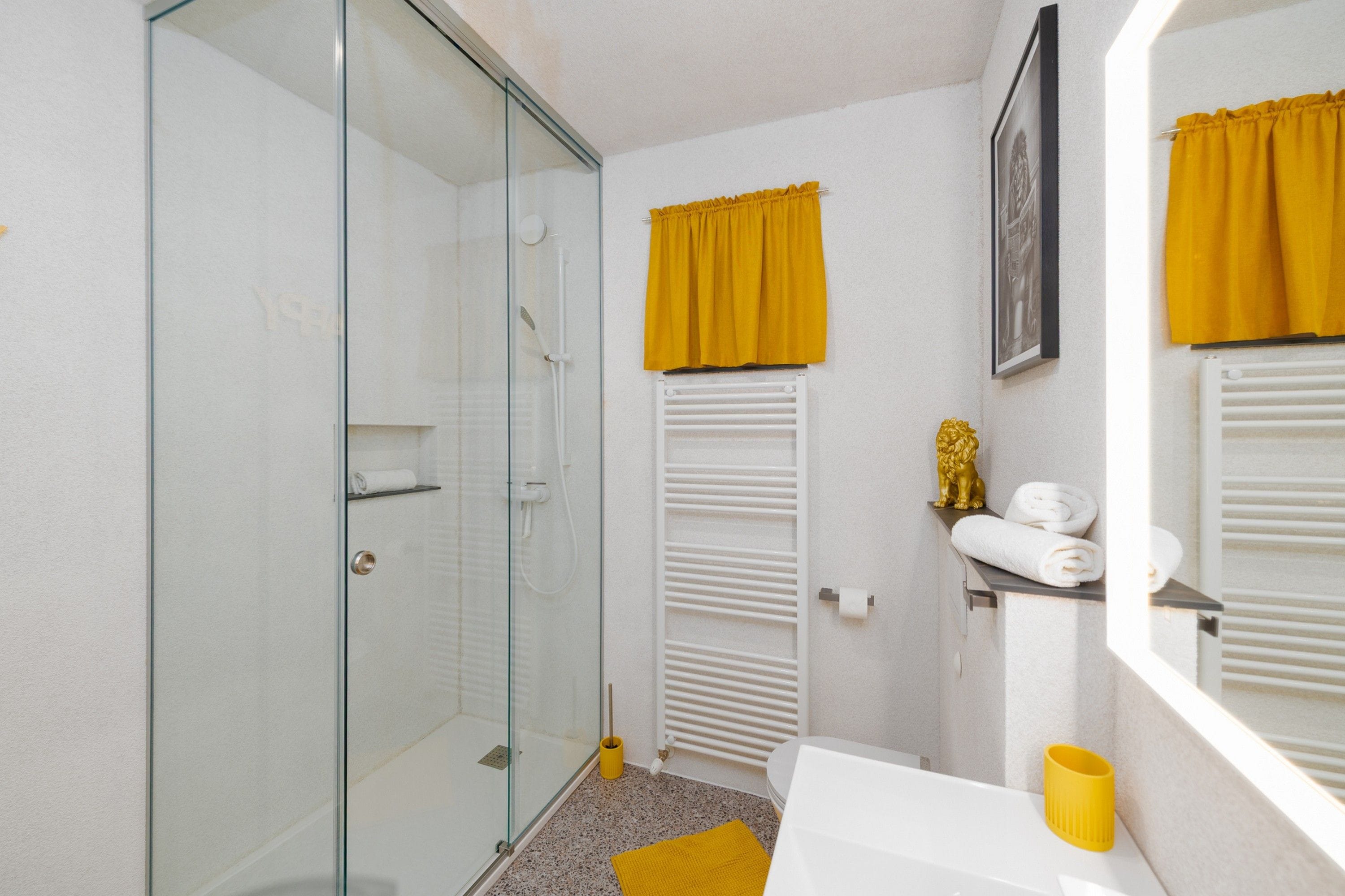 A bathroom with a glass shower enclosure, a white sink, a yellow towel, and a golden statue.