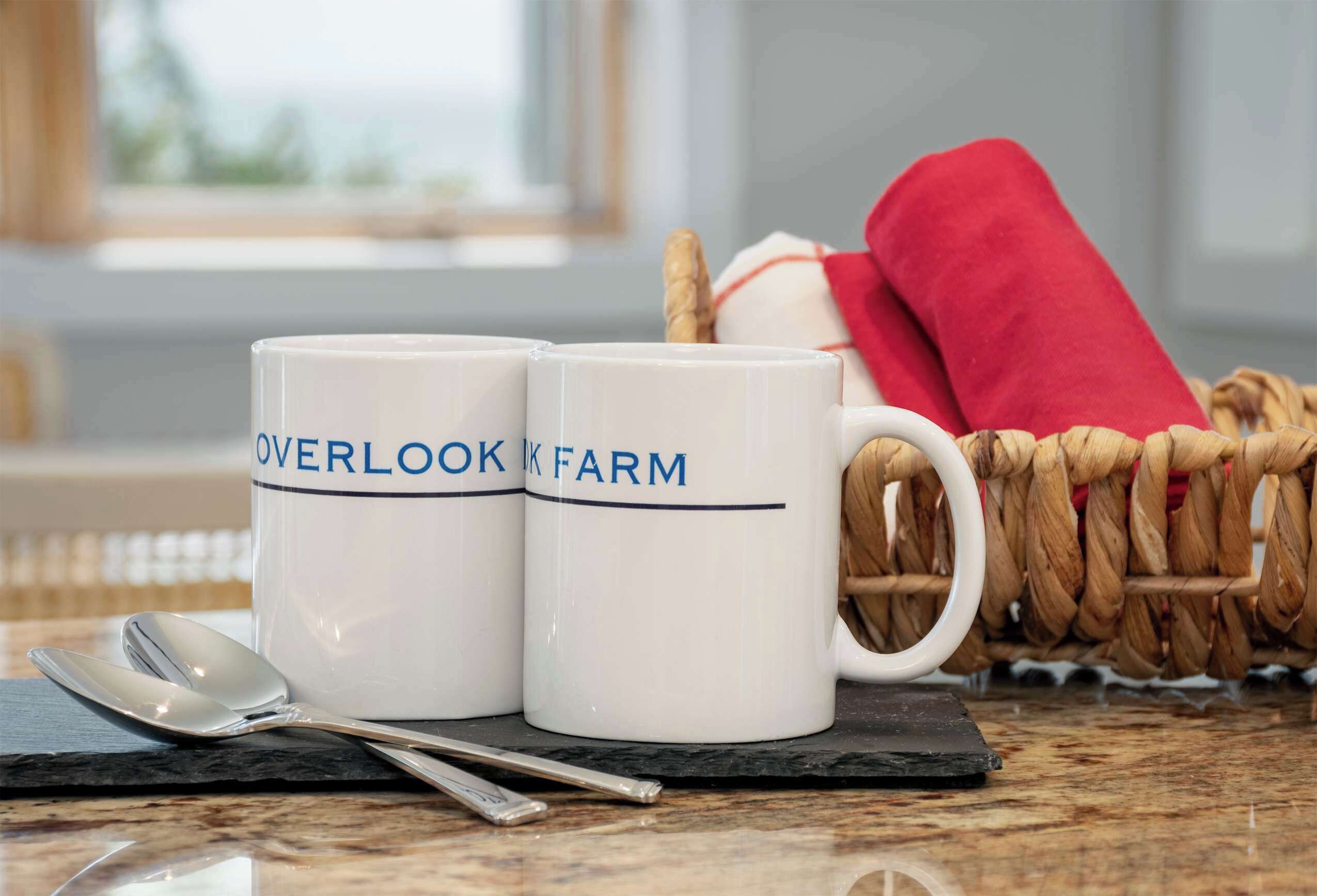 Two white mugs with "OVERLOOK FARM" text, a spoon, a knife, and a wicker basket with a red cloth.