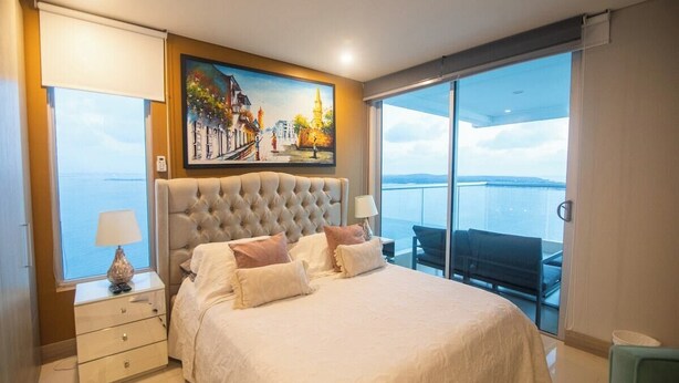 A bedroom with a large bed, a painting above the bed, a nightstand, a lamp, and a view of the sea through large windows.