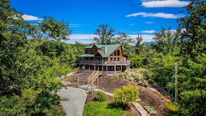 A two-story log cabin with a green roof, surrounded by dense forest, and a stone chimney.