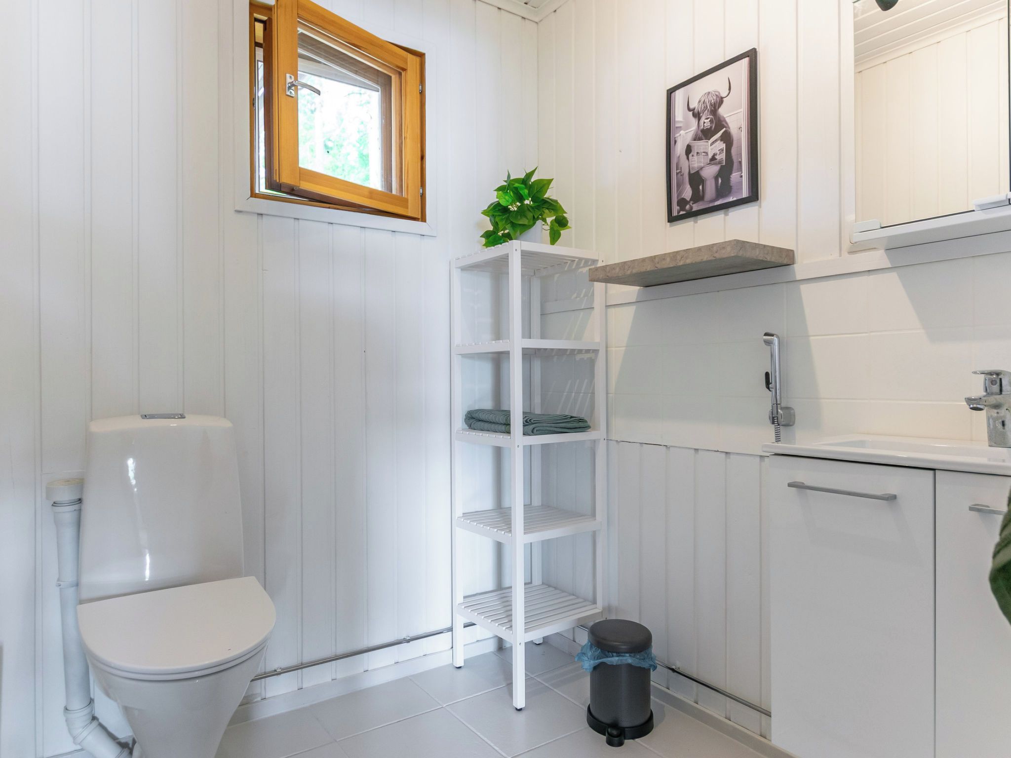 A bathroom with a toilet, a shelf unit, a trash bin, and a framed picture on the wall.