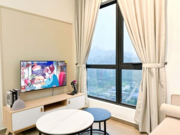 Ein Wohnzimmer mit Fernseher auf einem Holzständer, einem runden Tisch und Blick auf eine Stadtlandschaft durch ein großes Fenster mit Vorhängen.