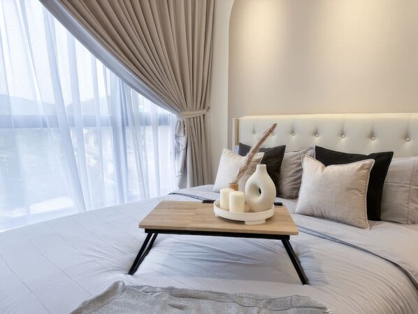 Une chambre moderne avec un grand lit, une table de chevet en bois ornée de bougies et une vue sur la mer à travers de fines rideaux.