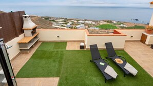 Eine Dachterrasse mit Grill, zwei Liegestühlen und Blick auf das Meer sowie entfernte Gebäude.