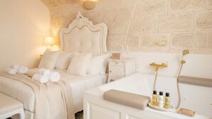 A hotel room with a stone wall, a bed with a white headboard, a bedside table with a lamp, and a small tray with toiletries. - Reale - Suite with Jacuzzi in the Heart of Monopoli (Monopoli)