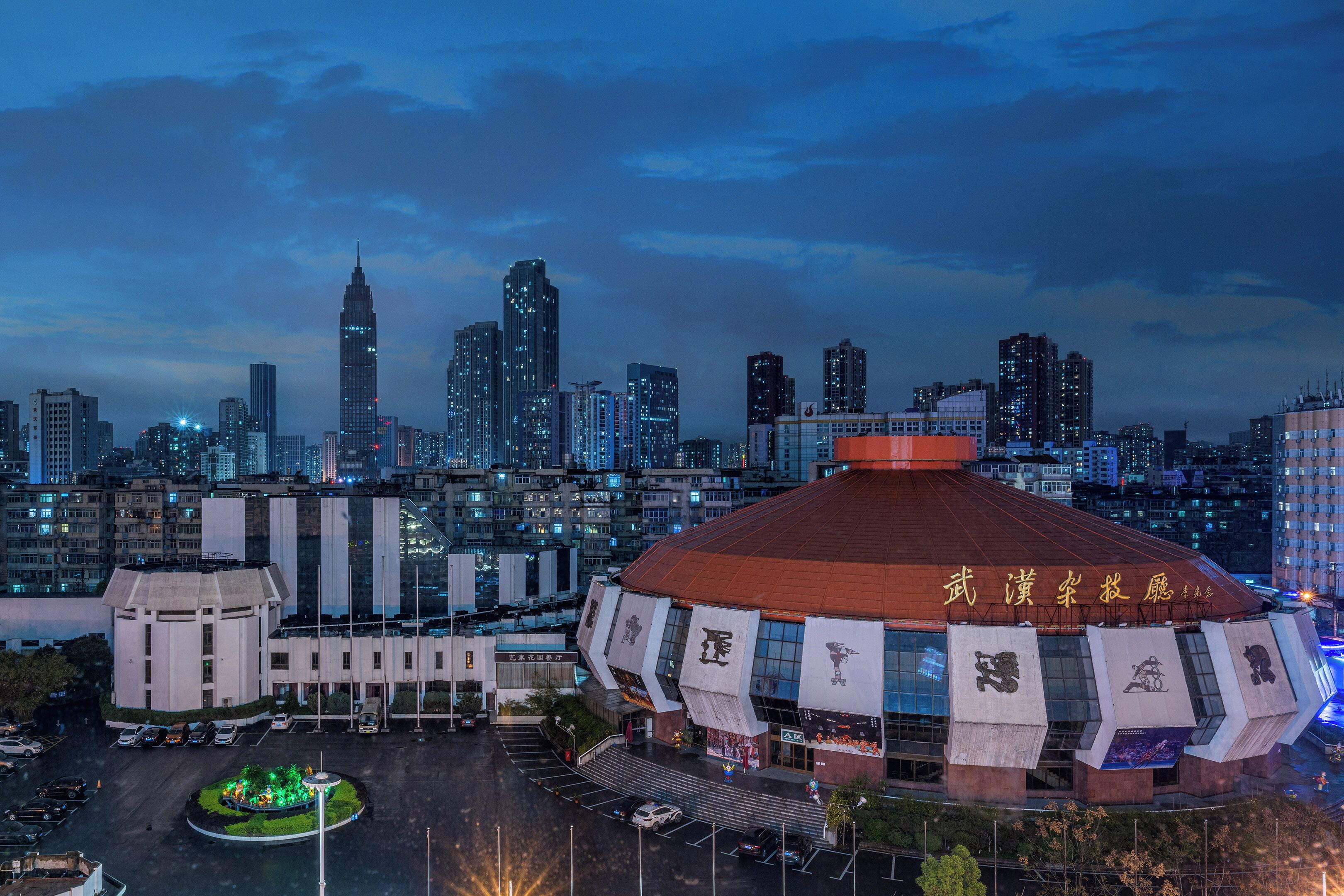 A large, uniquely shaped building with a red roof and multiple banners, surrounded by a cityscape with tall buildings and a parking area.