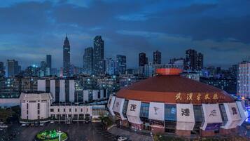 A large, uniquely shaped building with a red roof and multiple banners, surrounded by a cityscape with tall buildings and a parking area.