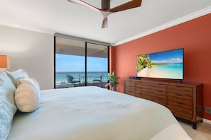 Un dormitorio con una cama grande, un televisor sobre una cómoda de madera, un ventanal corredero a un balcón y vista al océano.