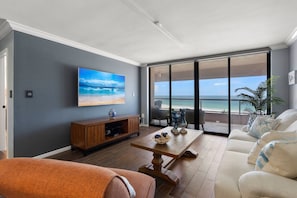 Una sala de estar con un televisor grande, una mesa de centro de madera, un sofá y vista al océano a través de un ventanal corredero.